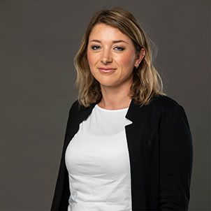 Portret Tamare Drmota v beli majici in črnem suknjiču, nasmejana pred temnim ozadjem